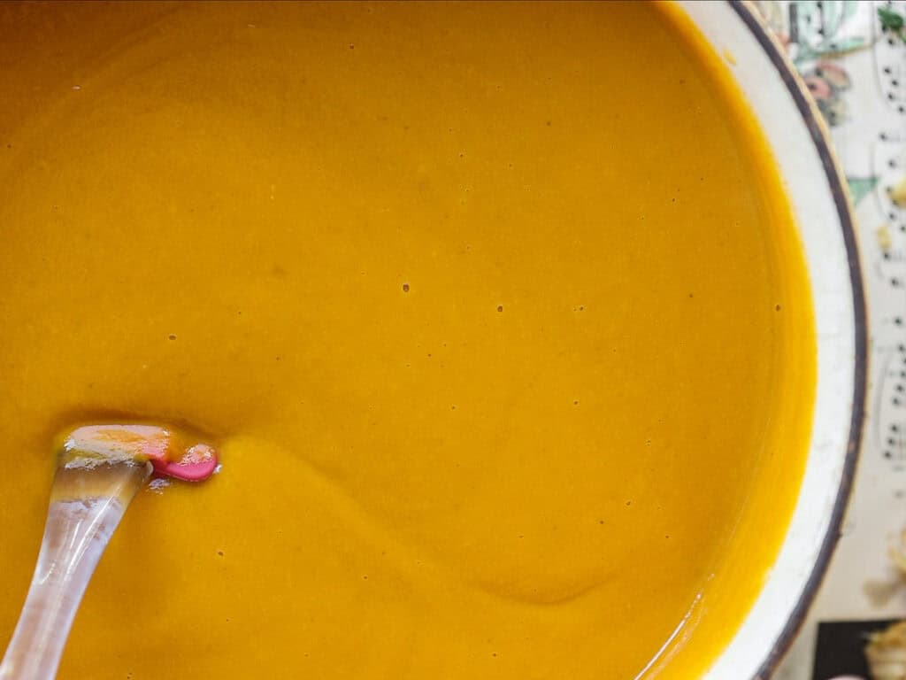 A close-up of a spoon dipped in a pot of creamy, orange-colored soup. The soup has a smooth texture, and the pot rests on a surface with a faint, patterned design visible on the edges.